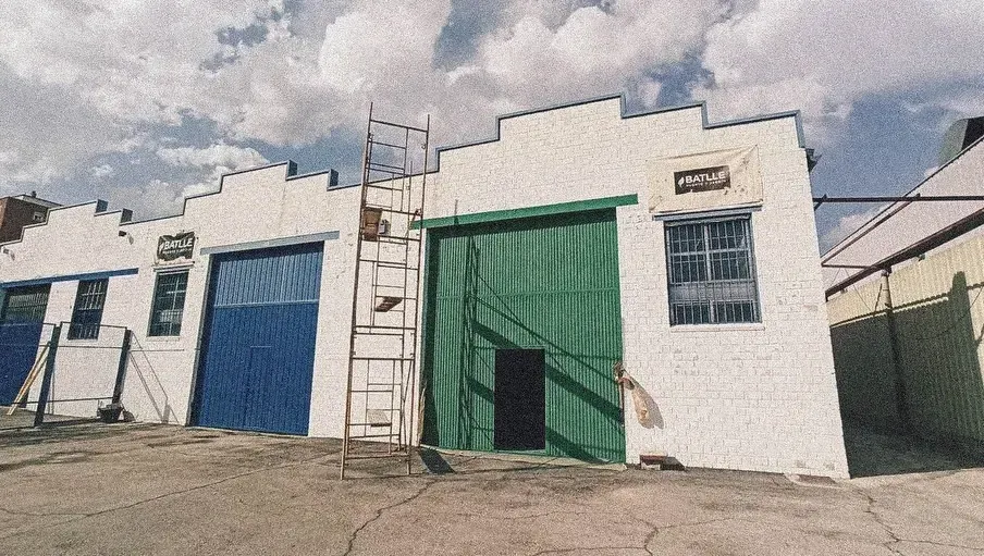 Hilera de edificios industriales blancos con puertas de garaje azules y verdes. Una escalera se alza contra uno de ellos.