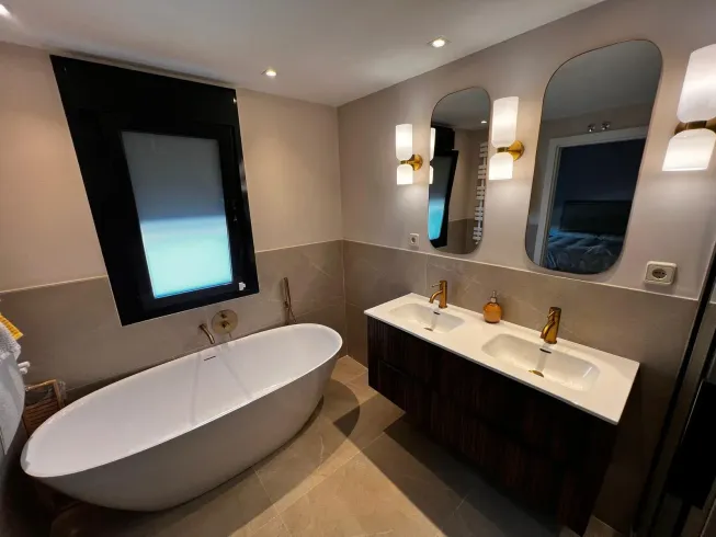 Baño moderno con bañera blanca, tocador doble con accesorios dorados y dos espejos arqueados.