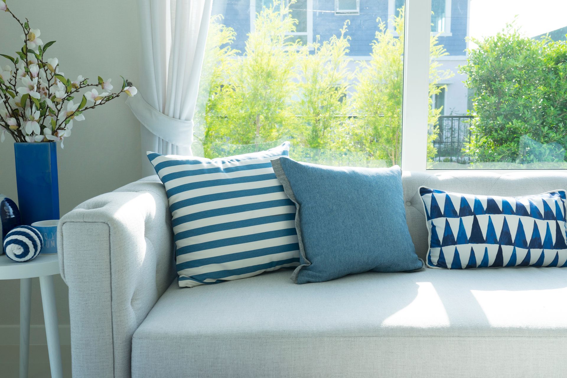 Weißes Sofa mit blau gestreiften, einfarbigen und gemusterten Kissen, daneben ein Tisch mit Blumen.