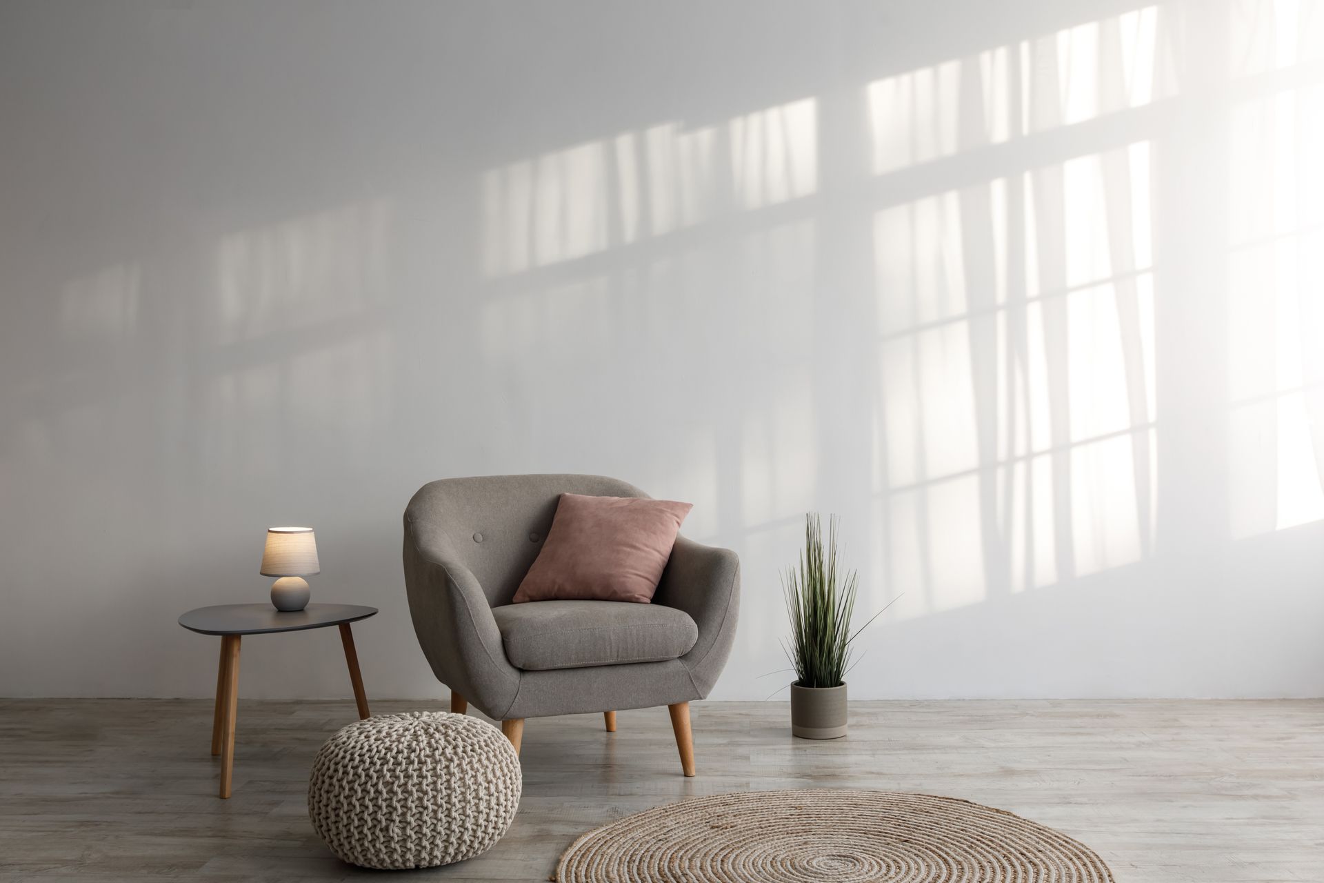 Grauer Sessel mit rosa Kissen, kleiner Tisch und Pflanze vor einer sonnenbeschienenen weißen Wand.