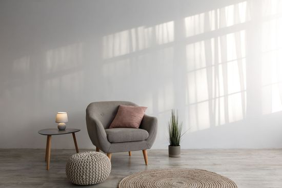Grauer Sessel mit rosa Kissen, kleiner Tisch und Pflanze vor einer sonnenbeschienenen weißen Wand.