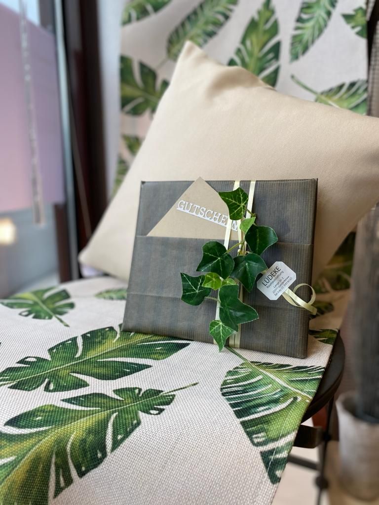 Ein Geschenk, verpackt in graues Papier, verziert mit Efeu und Schleifen, auf einer blattgemusterten Oberfläche, vor einem cremefarbenen Kissen.
