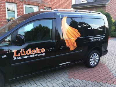 Schwarzer Lieferwagen mit dem Logo „Lüdeke Raumausstattung“ und orangefarbenem Vorhangmotiv, geparkt auf einer gepflasterten Auffahrt.