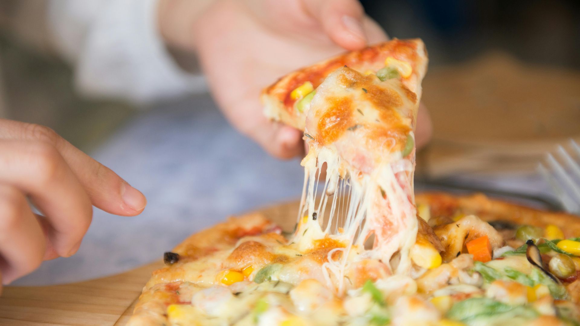 Persona levantando una porción de pizza con largas tiras de queso, desde una tabla de madera.