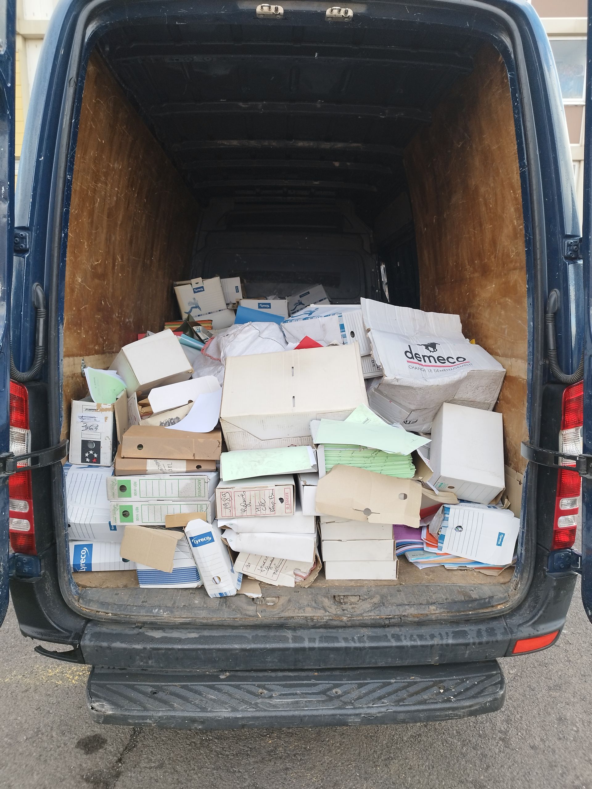 Les portes arrière d'une camionnette sont ouvertes, révélant un espace de chargement rempli de cartons empilés.