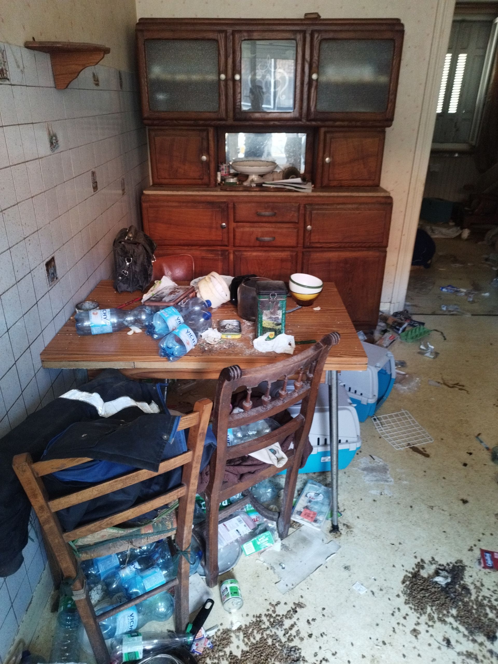 Une pièce encombrée avec un vaisselier en bois, une table recouverte de déchets et de bouteilles en plastique.