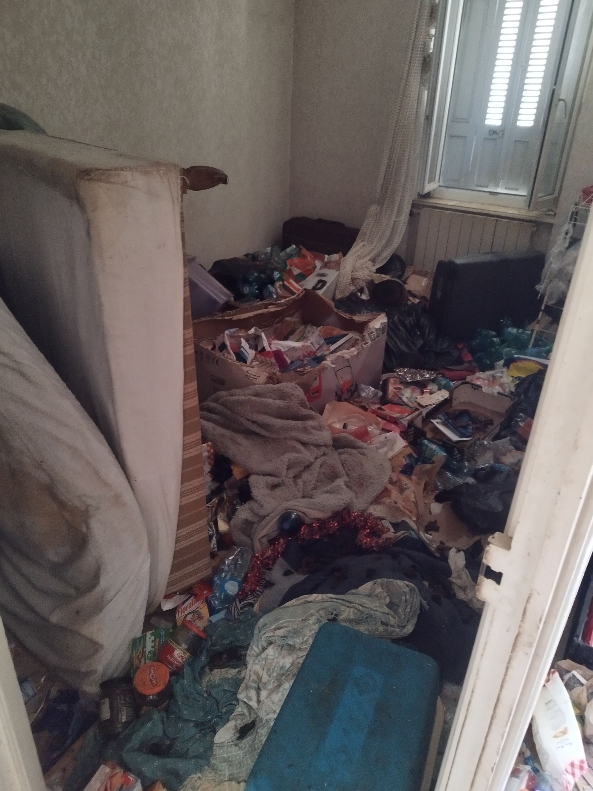 Une pièce encombrée et désorganisée, remplie d'objets ménagers éparpillés, d'emballages jetés et de matelas penchés.