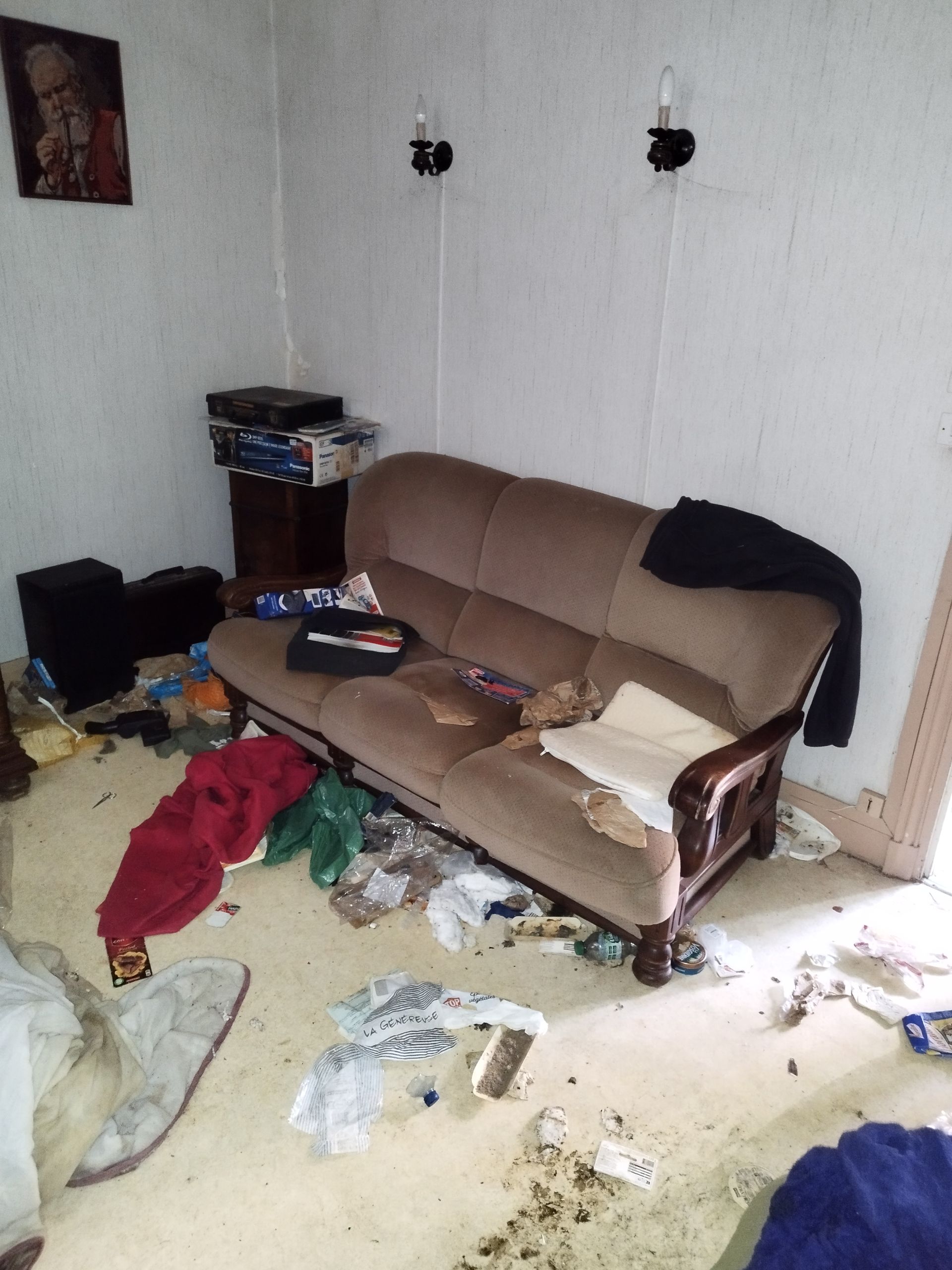 Un canapé marron est adossé à un mur blanc dans une pièce encombrée où jonchent le sol des détritus et des effets personnels.