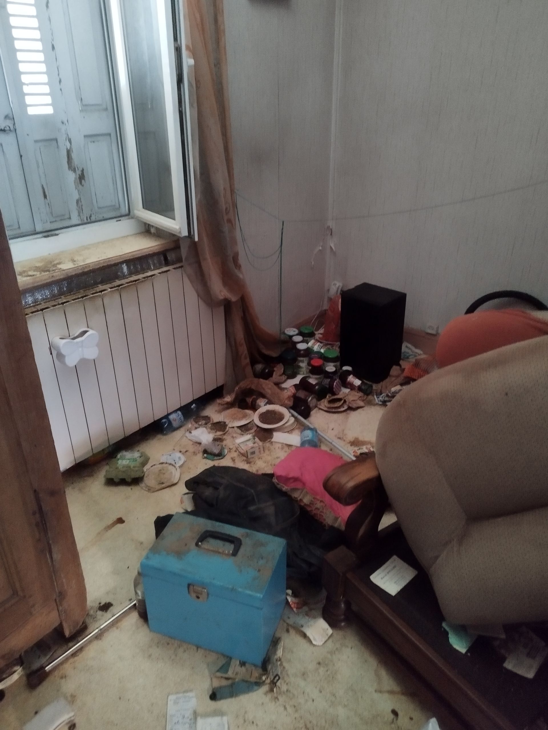 Une pièce encombrée avec une boîte à outils bleue, un fauteuil marron et des débris éparpillés sur le sol..