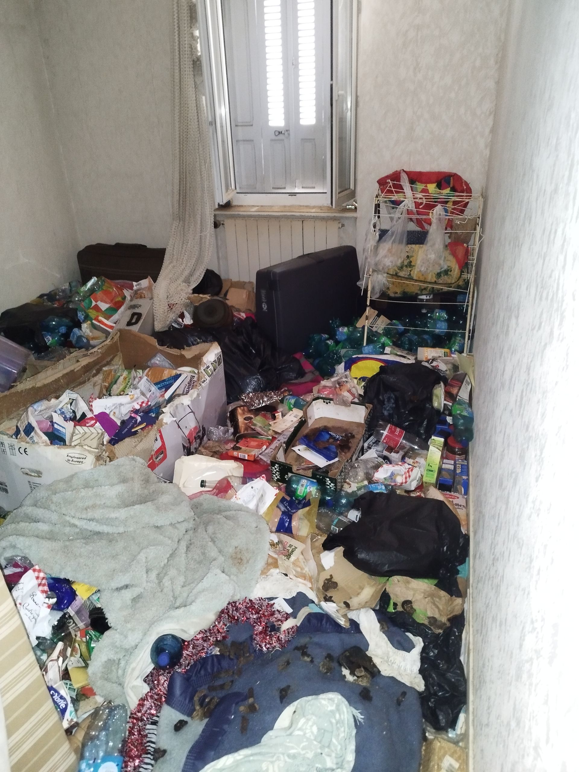 Une pièce étroite et encombrée, remplie de sacs, de cartons, de déchets et d'objets personnels éparpillés sur le sol.