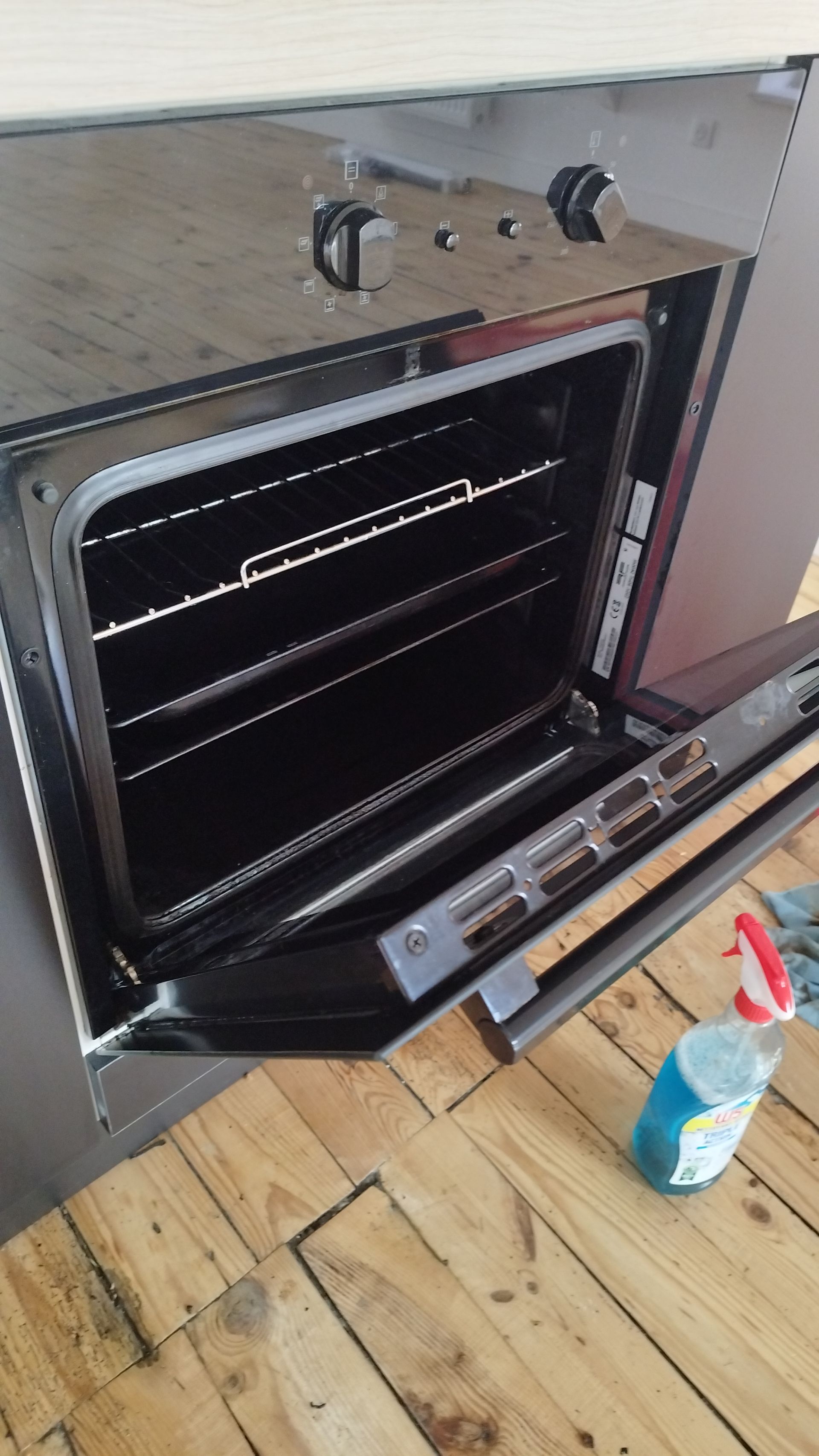 Four mural noir ouvert dans une pièce au parquet clair, avec un flacon de nettoyant aérosol posé à proximité.