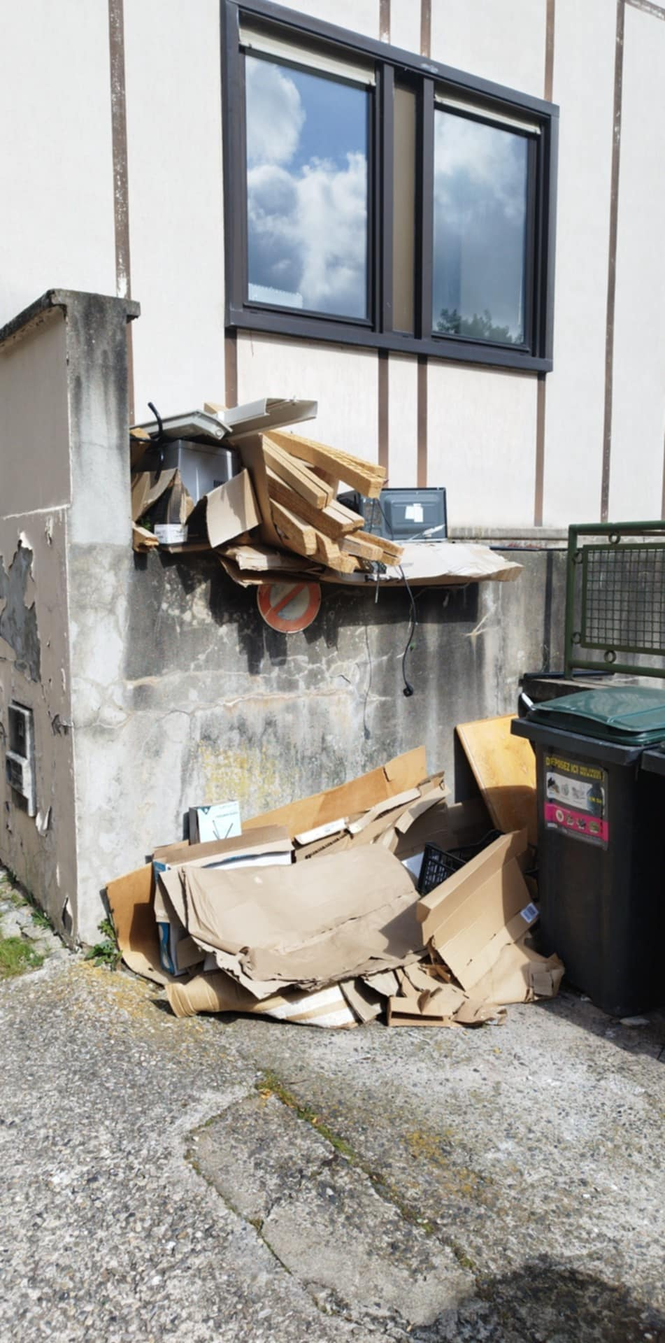 Des cartons et des objets divers empilés contre un mur de béton, sous une fenêtre où se trouve une poubelle verte.