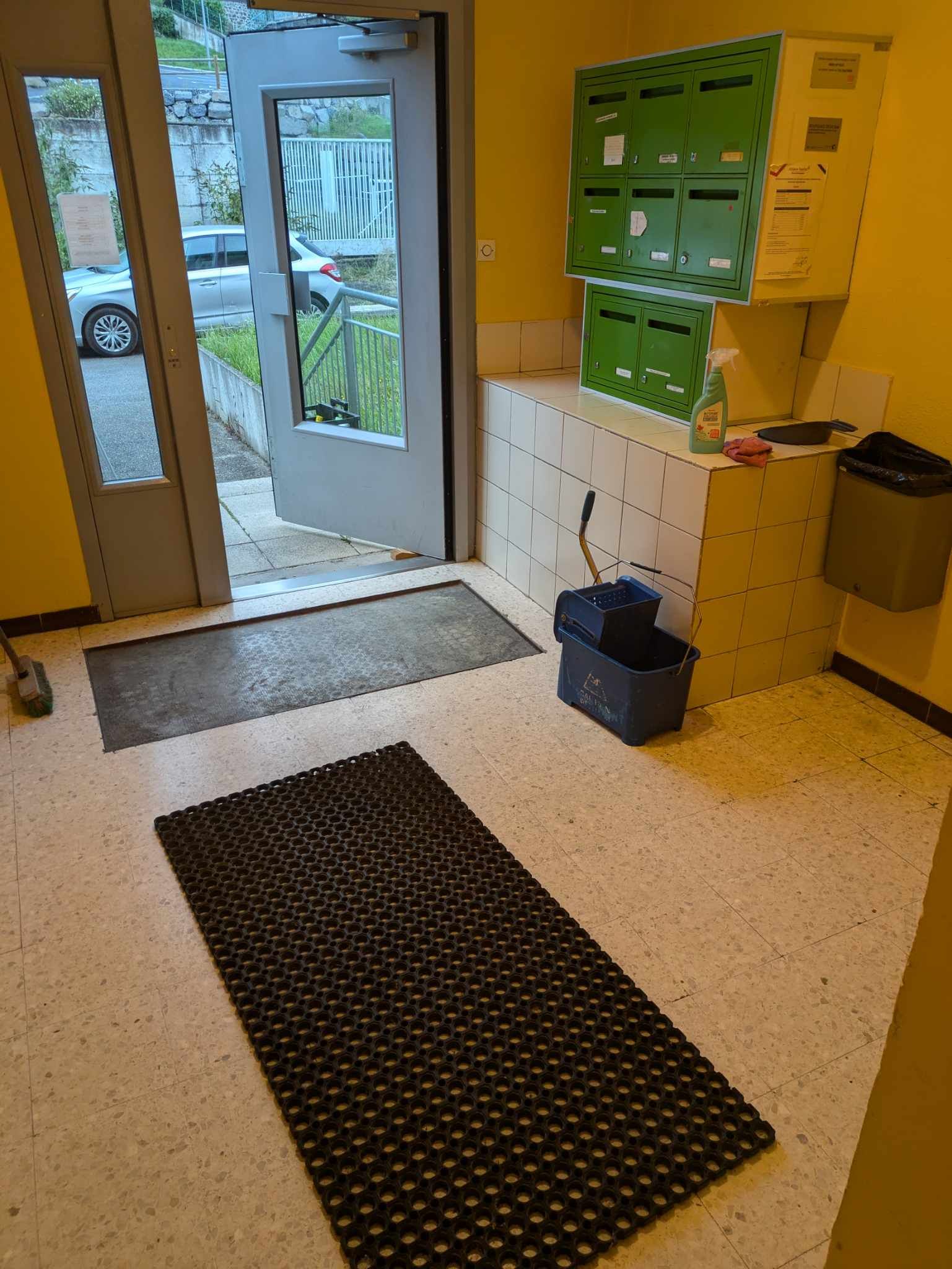Un hall d'entrée aux murs jaunes avec des boîtes aux lettres et du matériel de nettoyage.