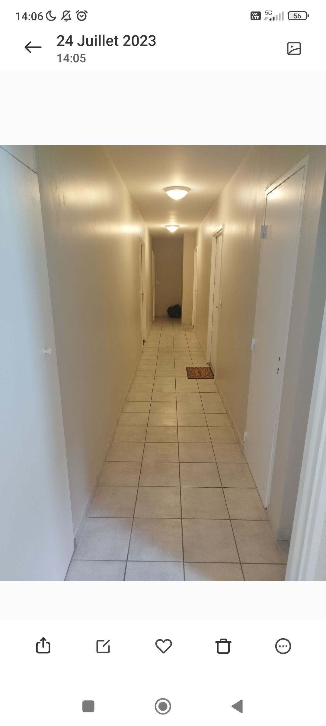 Un long couloir étroit aux murs blancs, au sol carrelé et éclairé par deux plafonniers, menant à une porte au fond.