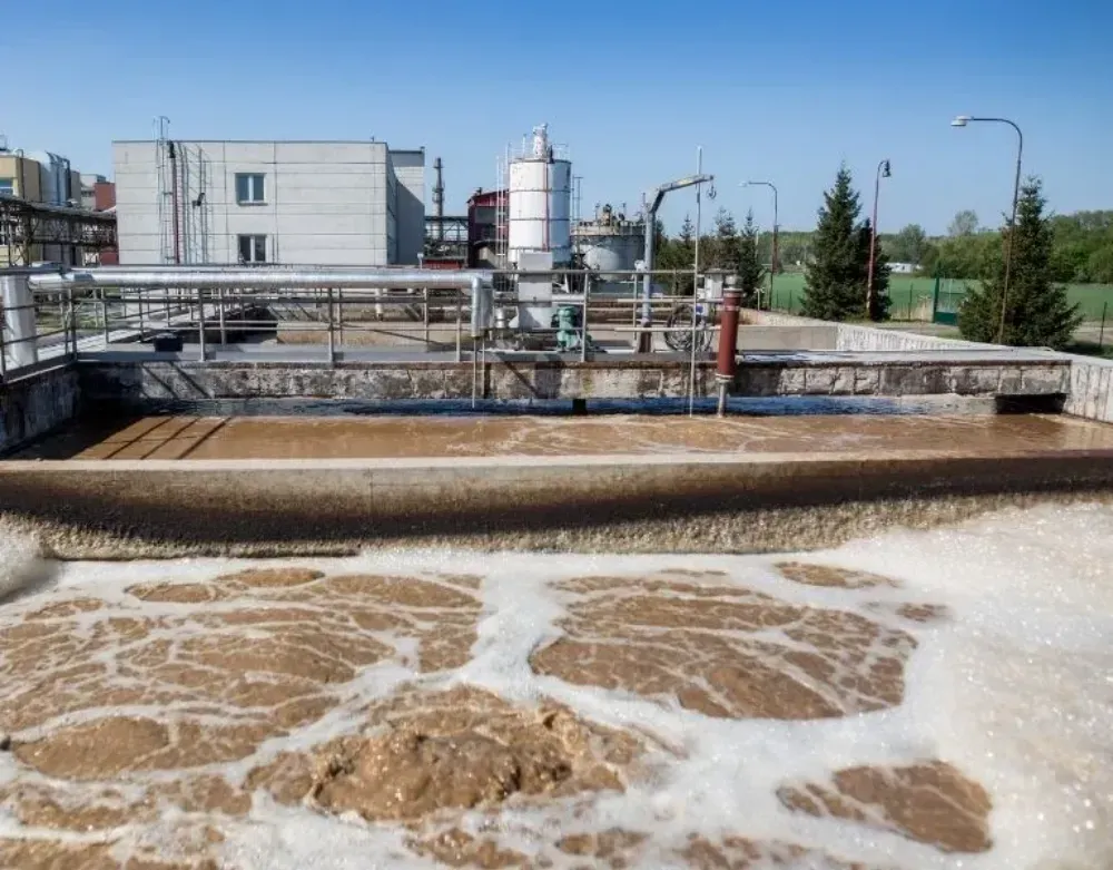 Tanques de tratamiento de aguas residuales con agua marrón espumosa y edificios industriales al fondo