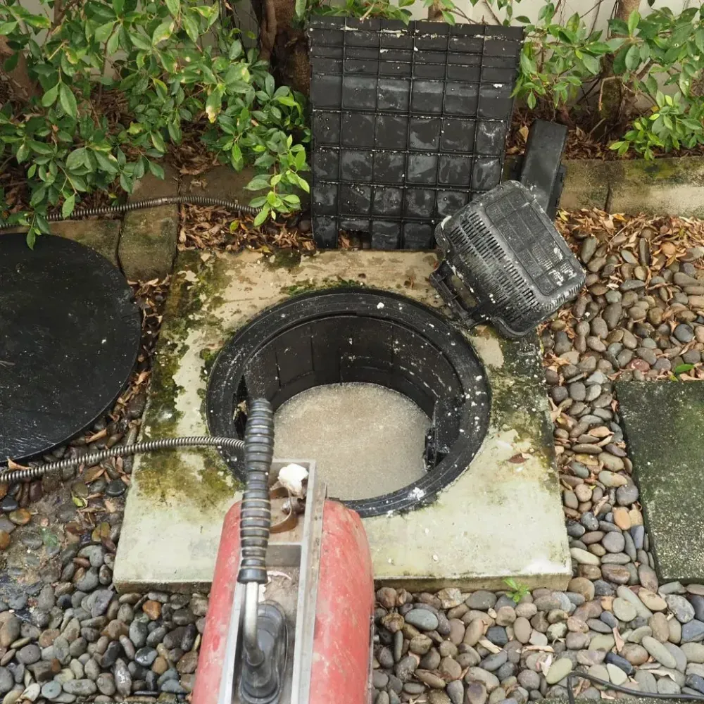 Abertura circular para acceder a los servicios en un jardín de grava, con una manguera y componentes de plástico negro cerca.