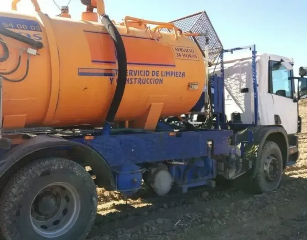 Camión cisterna de vacío naranja con chasis azul aparcado al aire libre sobre terreno irregular