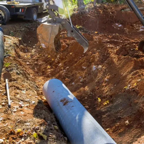 Una excavadora cavando una zanja alrededor de una tubería gris grande en una obra de construcción con tierra.