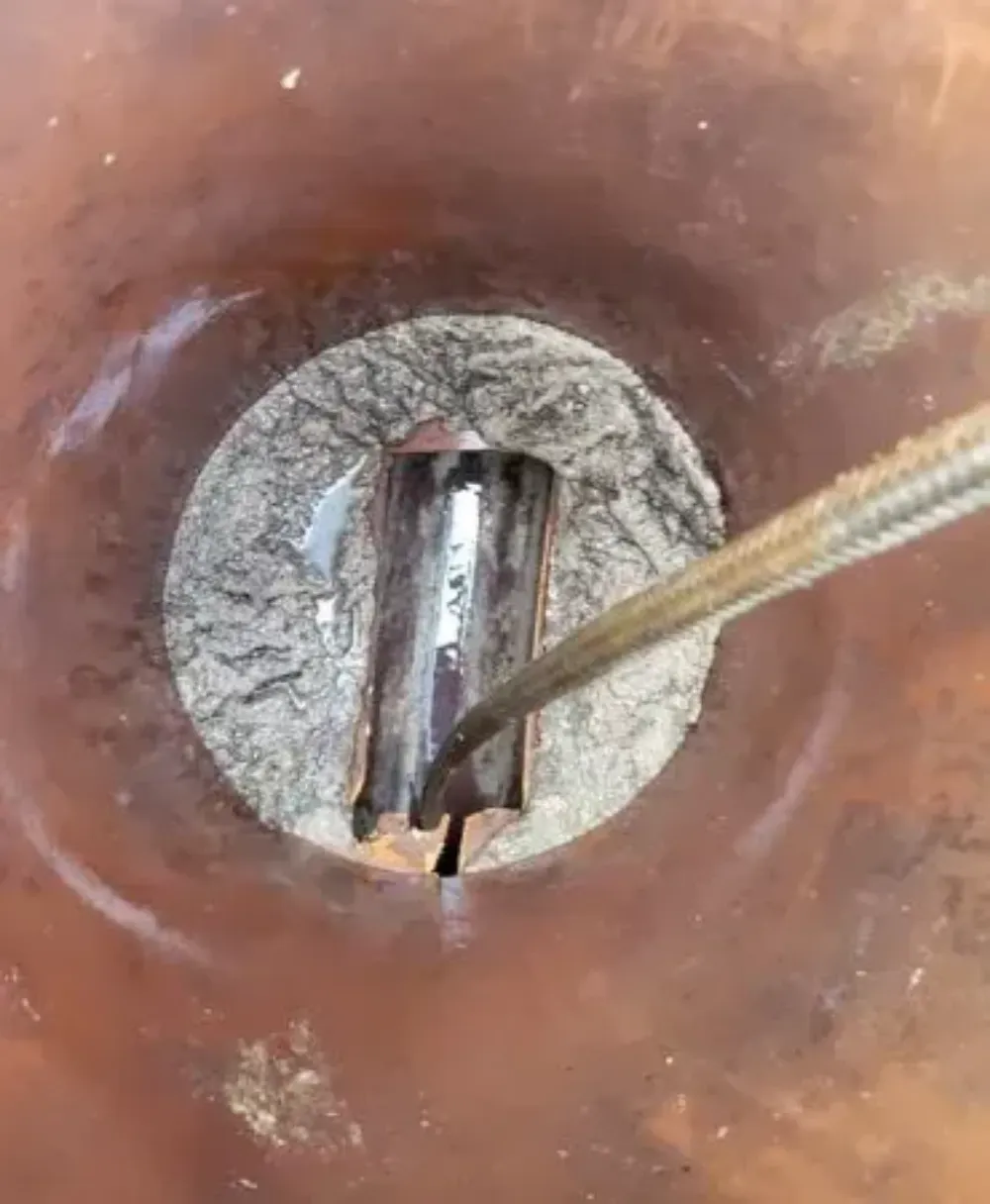 Primer plano de un desagüe metálico corroído, con un destornillador apuntando a la grieta del centro.