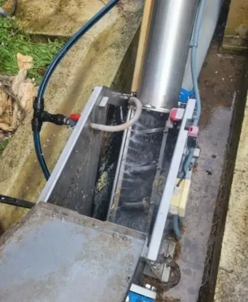 Bomba de agua metálica y tuberías en una zanja estrecha, con agua corriendo y terreno embarrado