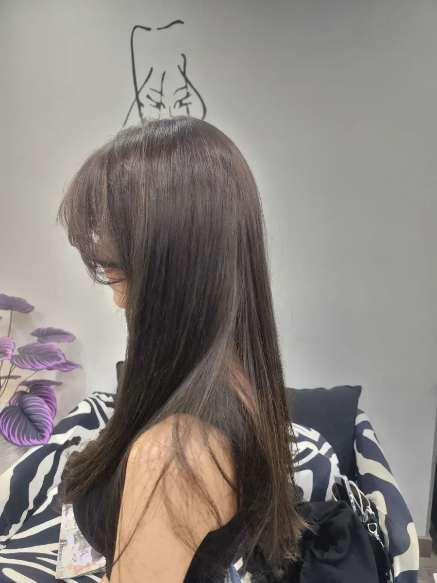 Perfil de una mujer con cabello largo castaño en una habitación con una planta de flores moradas.