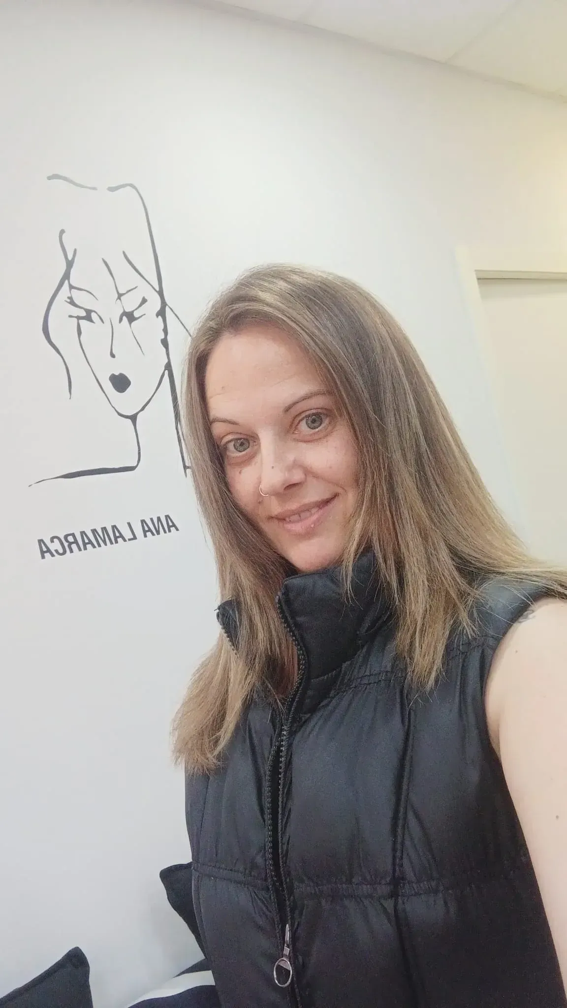 Mujer con chaleco negro tomándose una selfie en el interior de una casa, junto a una pared con un dibujo lineal.