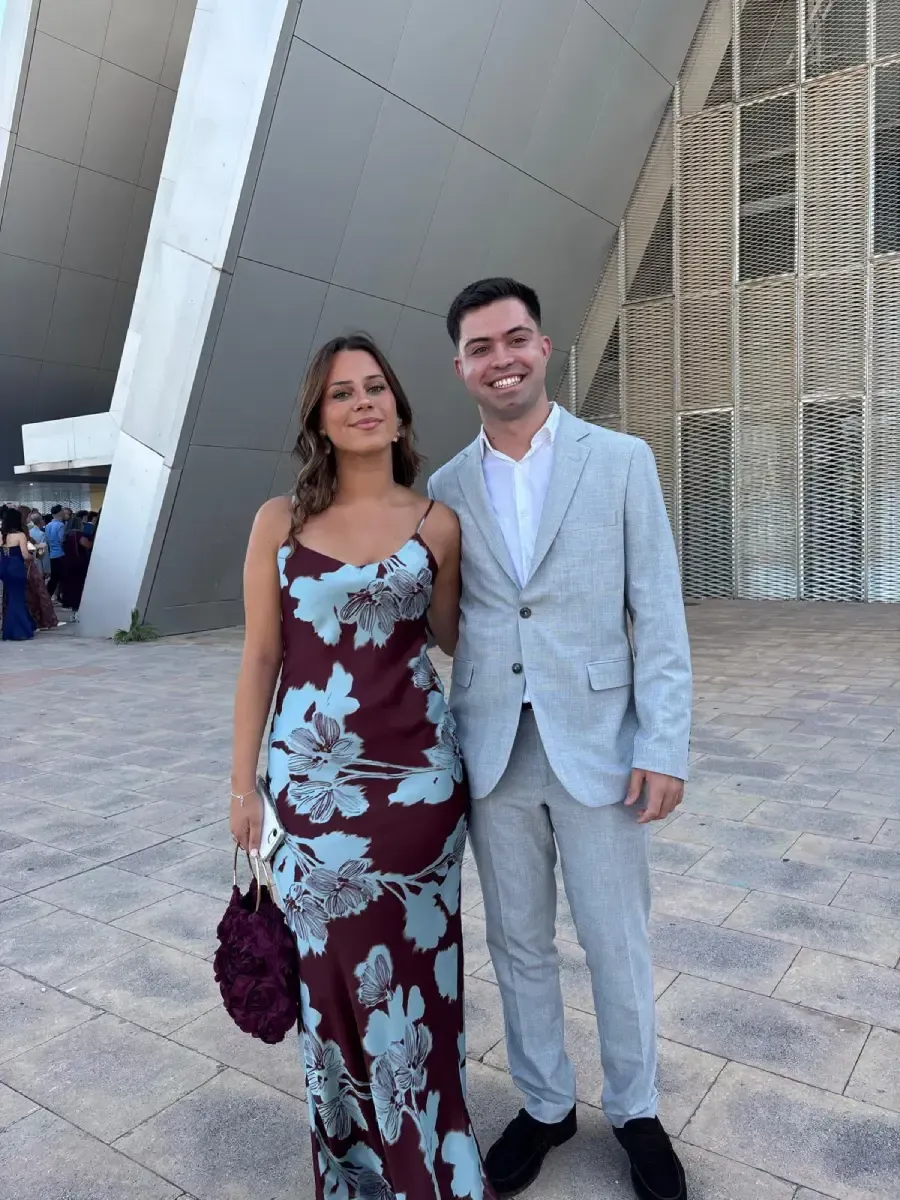 Dos personas posando frente a un edificio moderno, vestidas con atuendos formales, sonriendo a la cámara.