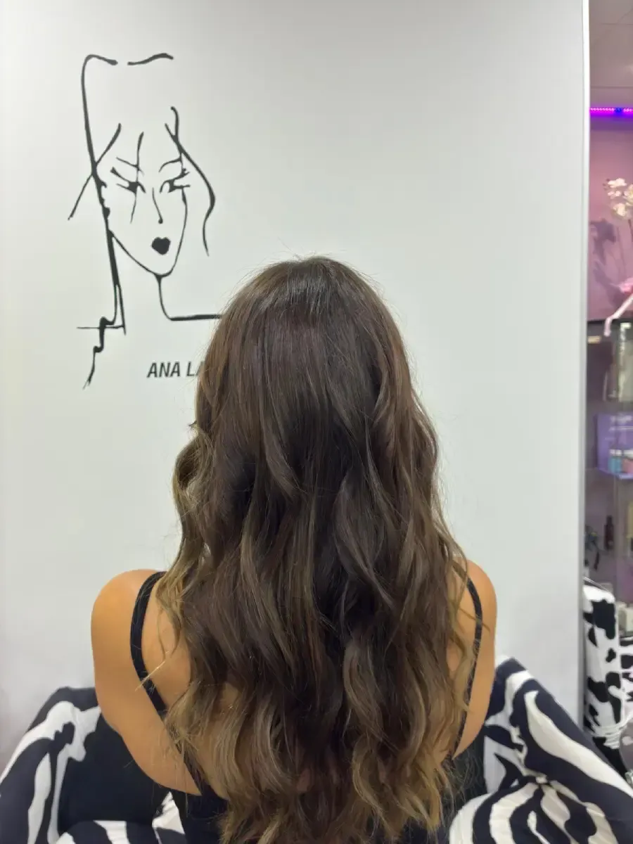 Mujer con cabello castaño ondulado vista de espaldas, frente a una ilustración en la pared de un salón luminoso.