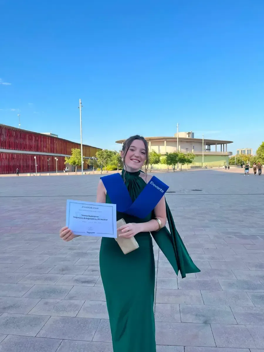 Mujer sosteniendo un certificado al aire libre en un campus soleado, vestida con un vestido verde