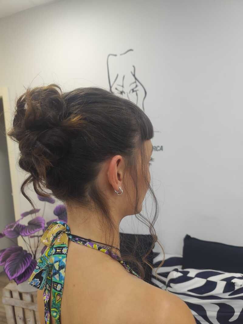 Mujer de cabello oscuro recogido en un moño, vista desde atrás, con una prenda colorida en un interior y frente a una pared blanca.