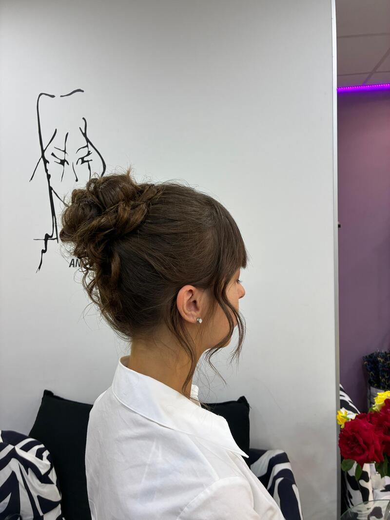 Perfil de una mujer con un moño rizado y una camisa blanca, de pie en el interior junto a una pared con un dibujo.