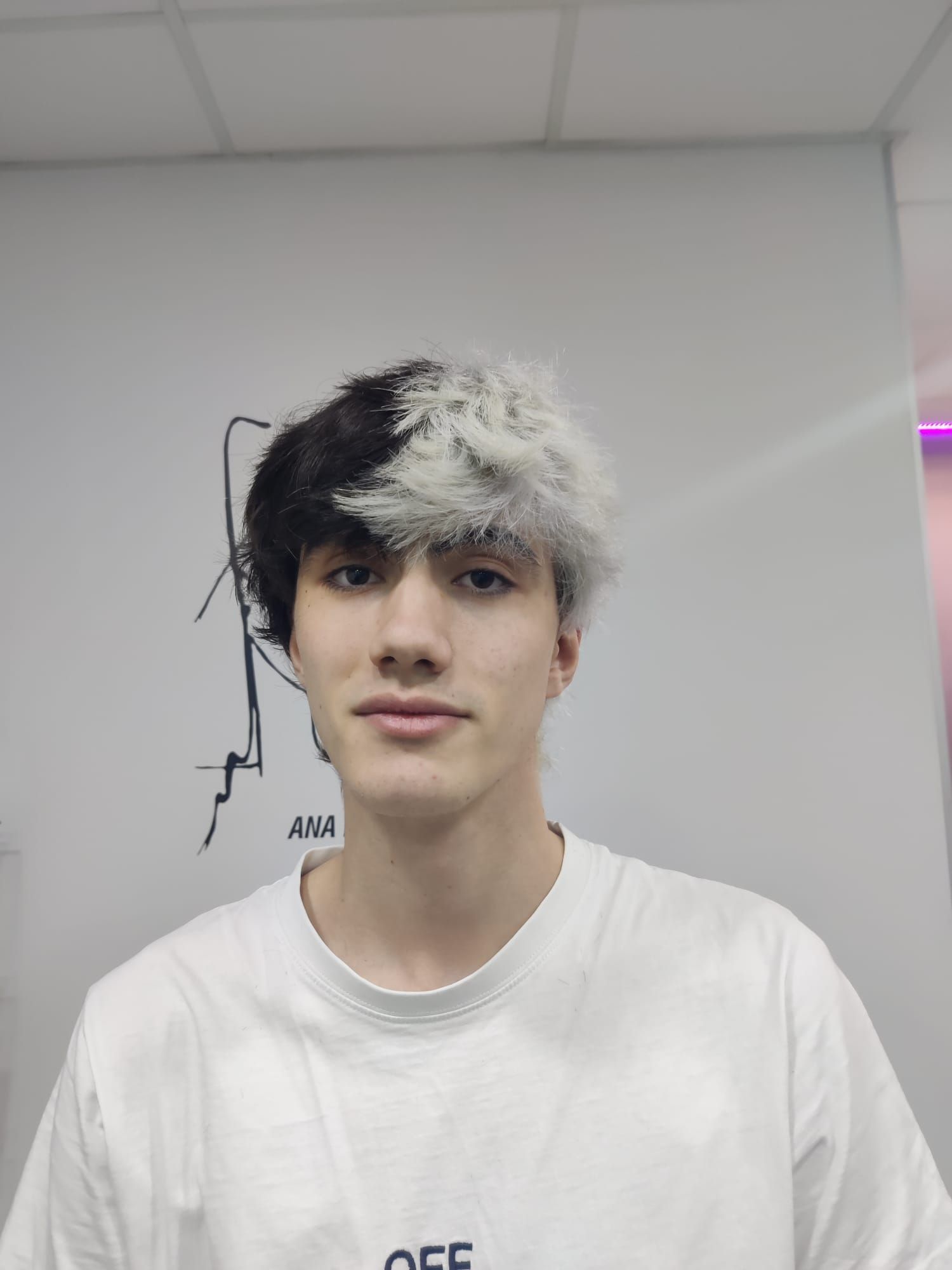 Persona con el pelo blanco y negro, con una camiseta blanca, de pie en el interior frente a una pared lisa.