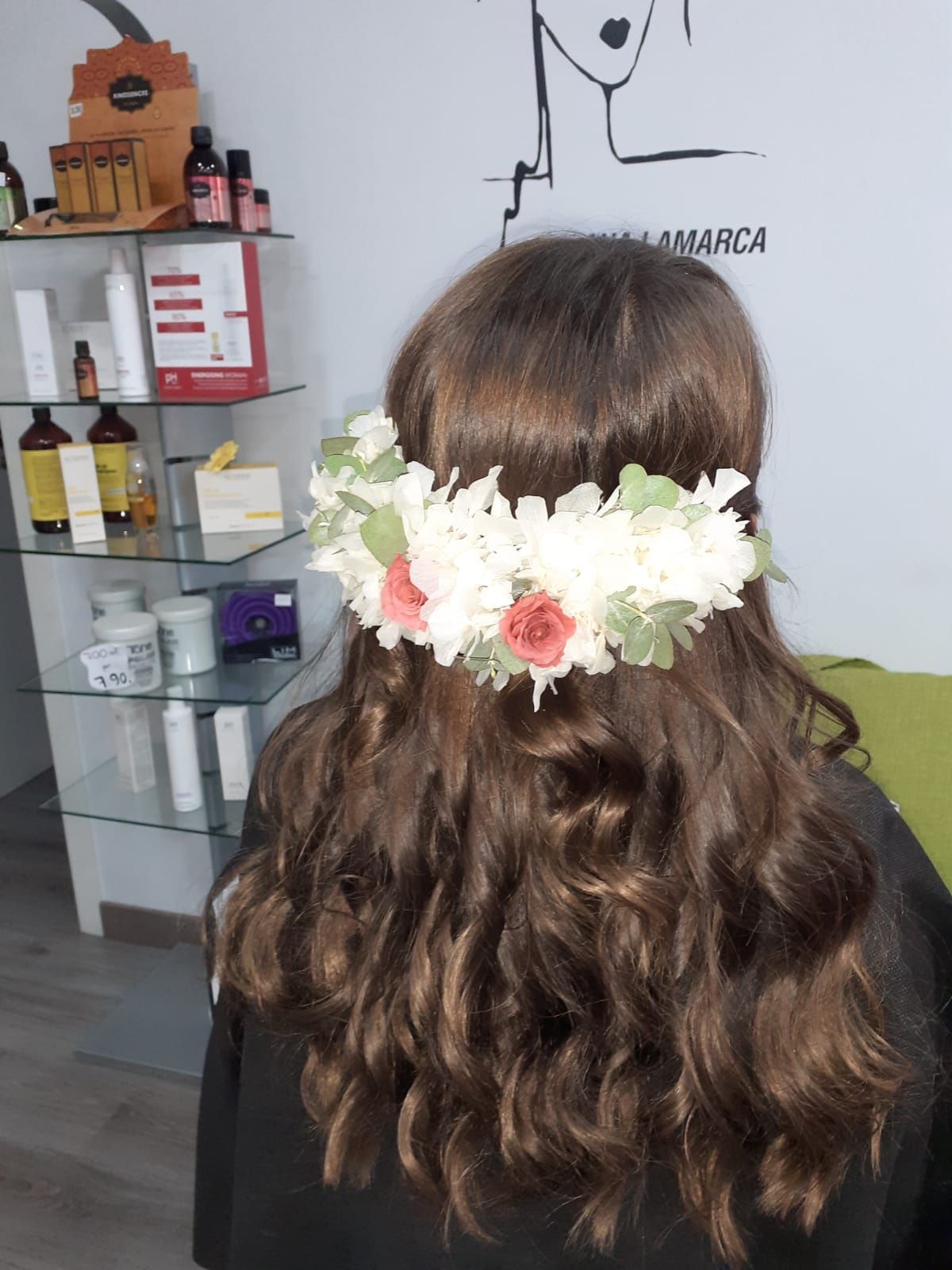 Persona con el pelo castaño ondulado que lleva una corona de flores blancas, vista desde atrás en un salón de belleza.