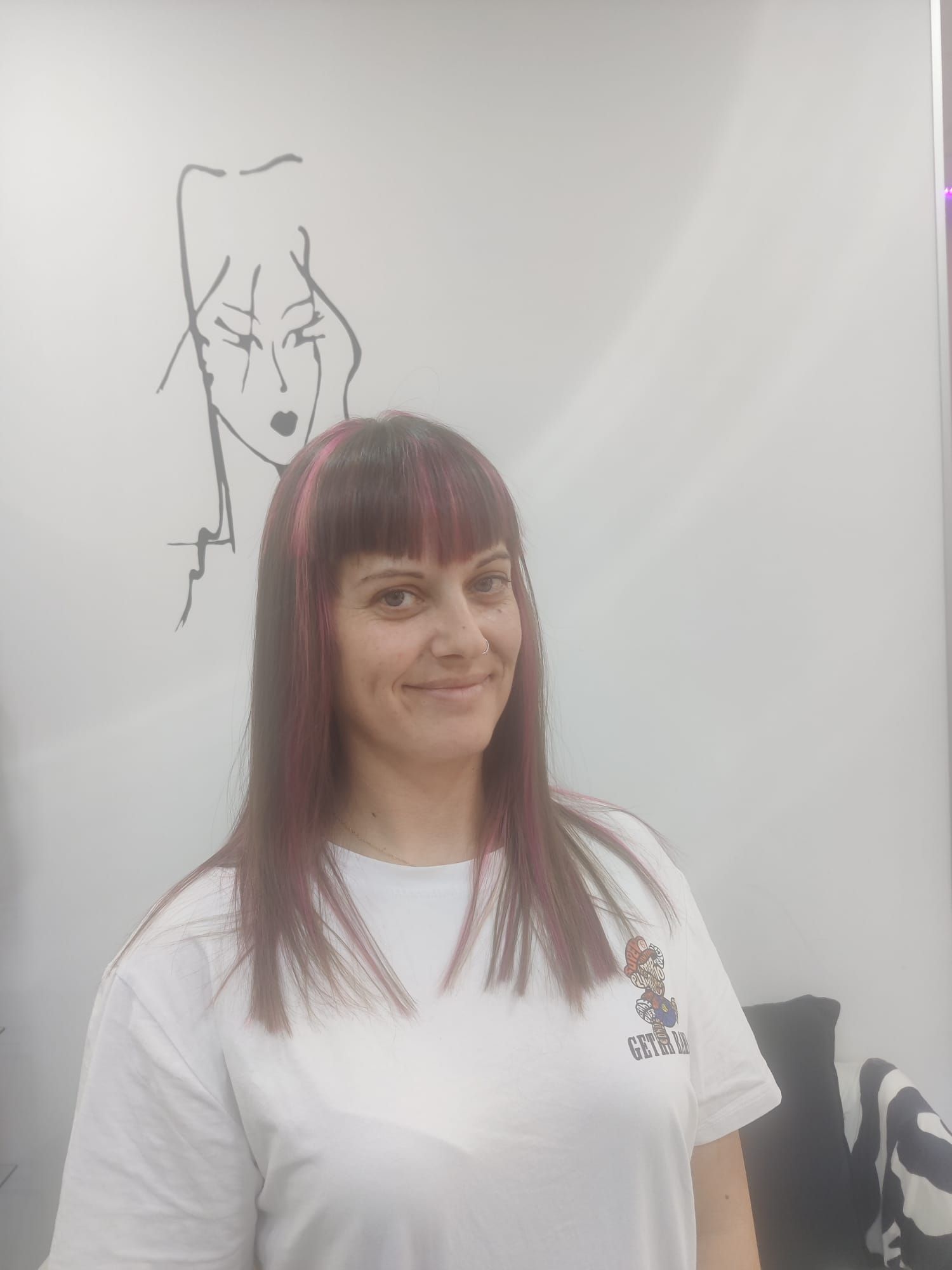 Mujer de cabello largo castaño rojizo, sonriendo con una camiseta blanca y de pie frente a una pizarra con un dibujo.