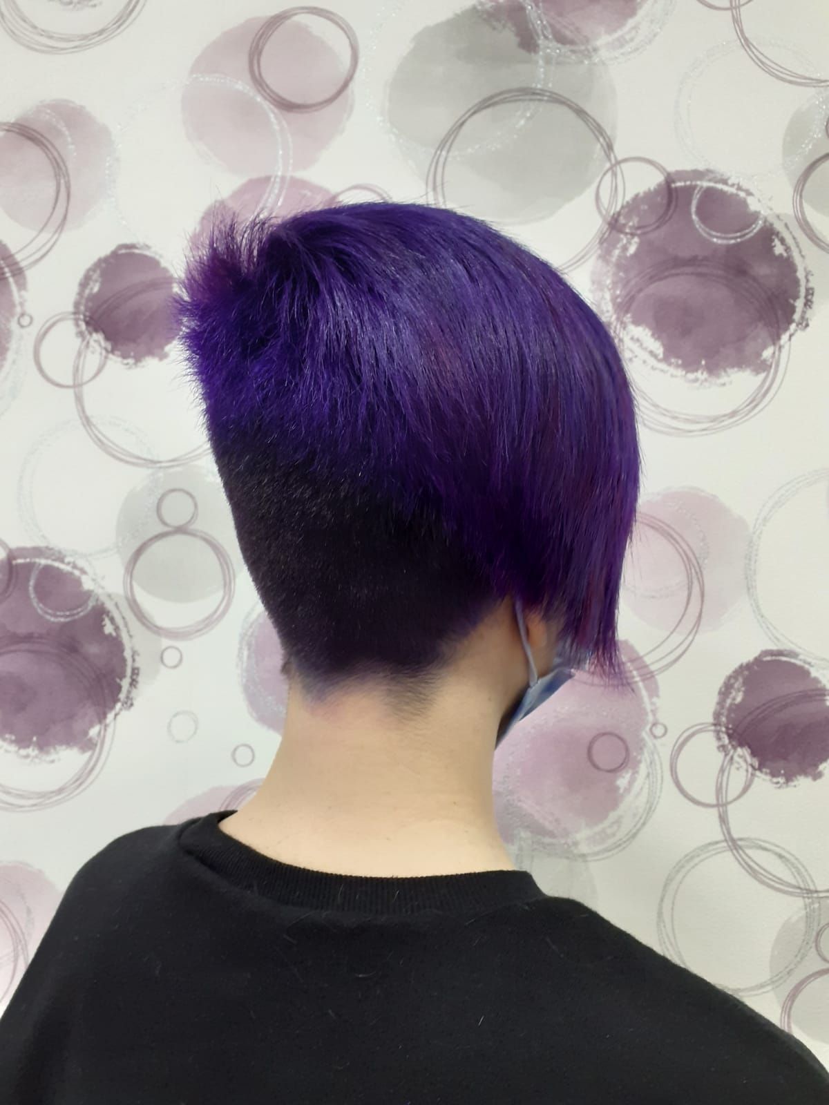 Vista trasera de un cabello corto y morado con un corte rapado a los lados, sobre una pared clara con estampado