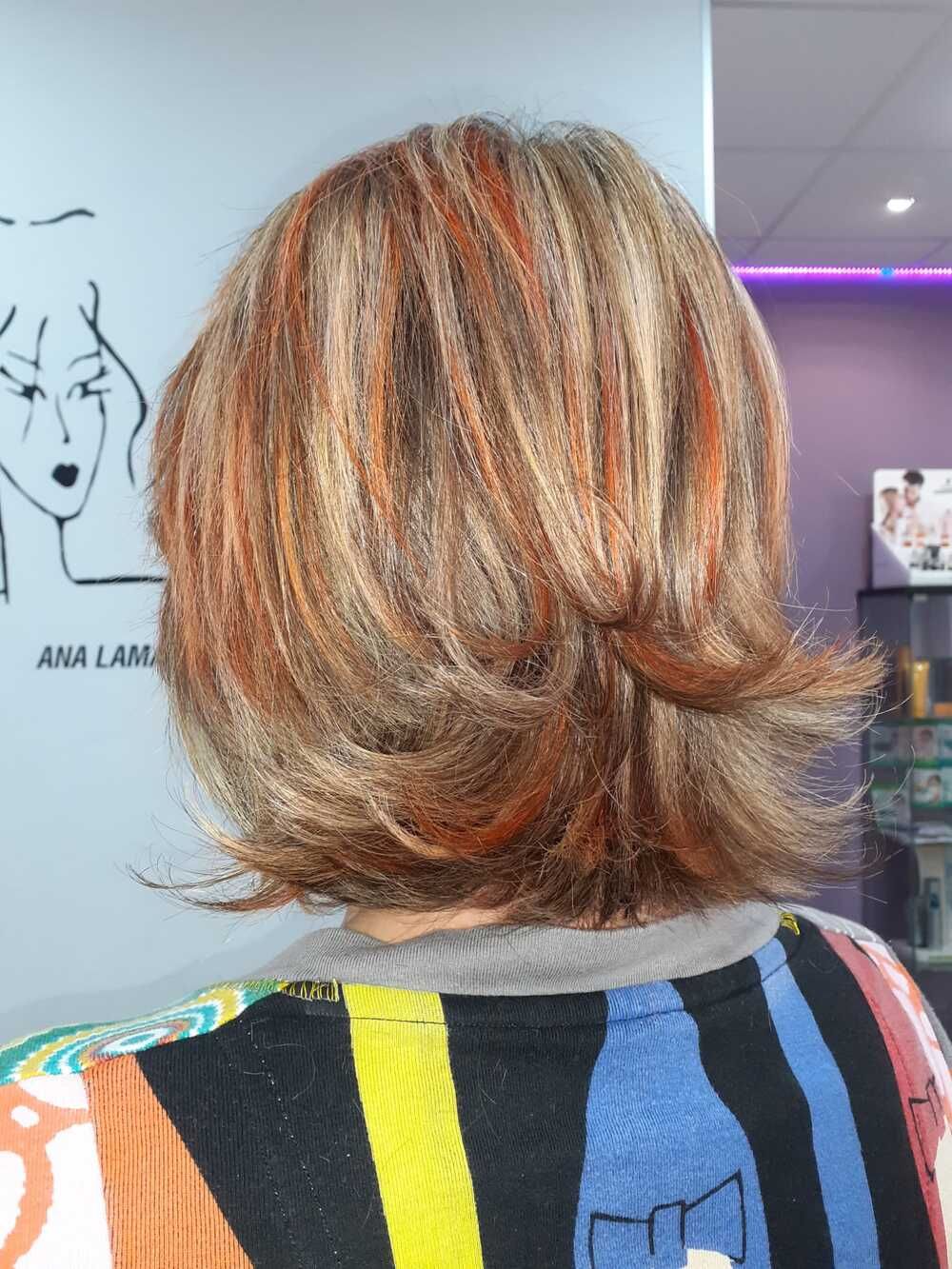 Vista trasera de una persona con el pelo corto, a capas, rubio y cobrizo en una peluquería, con una camiseta de rayas de colores