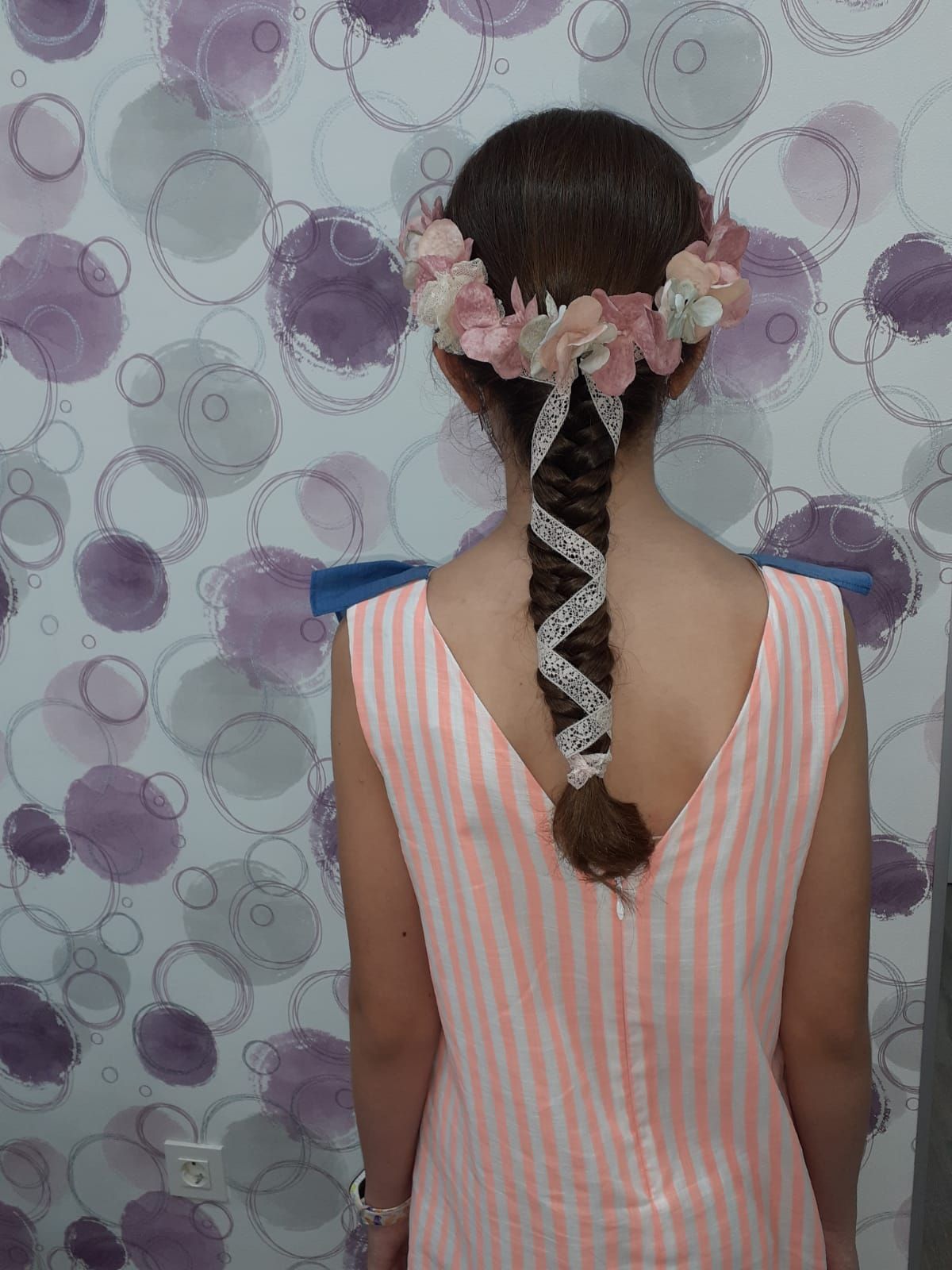 Chica con el pelo trenzado y una diadema floral, vista de espaldas frente a una pared estampada.