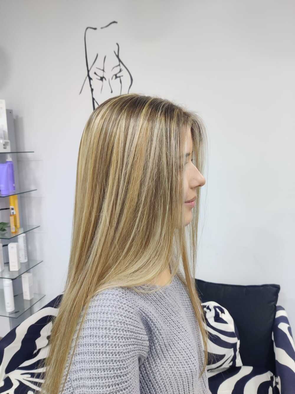 Vista de perfil de una mujer rubia de cabello liso sentada en una silla de peluquería, con un suéter gris.