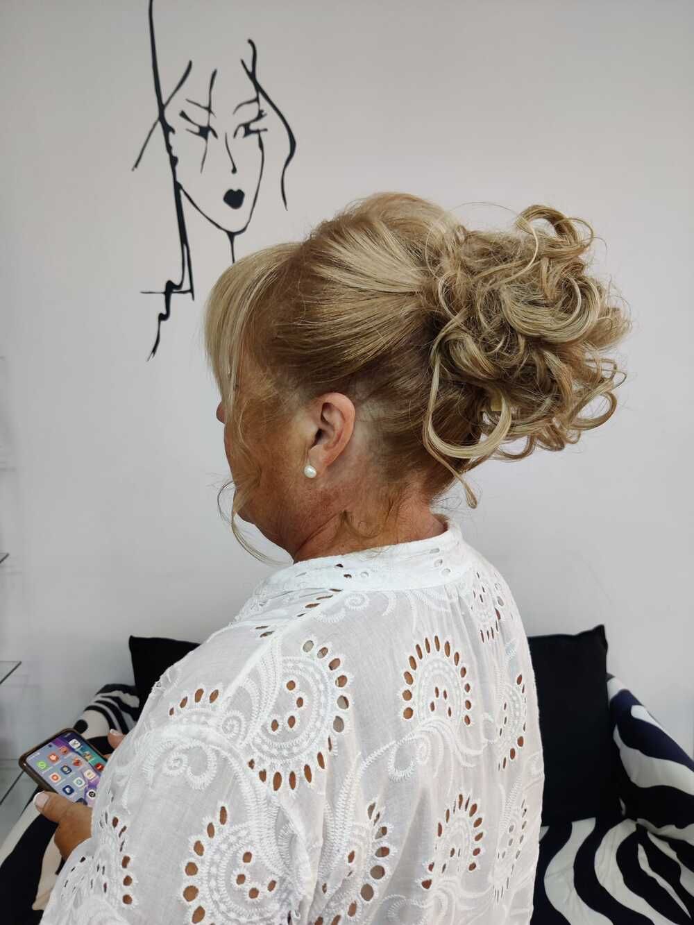 Mujer rubia con una blusa blanca calada, mirando un dibujo en la pared mientras sostiene un teléfono.