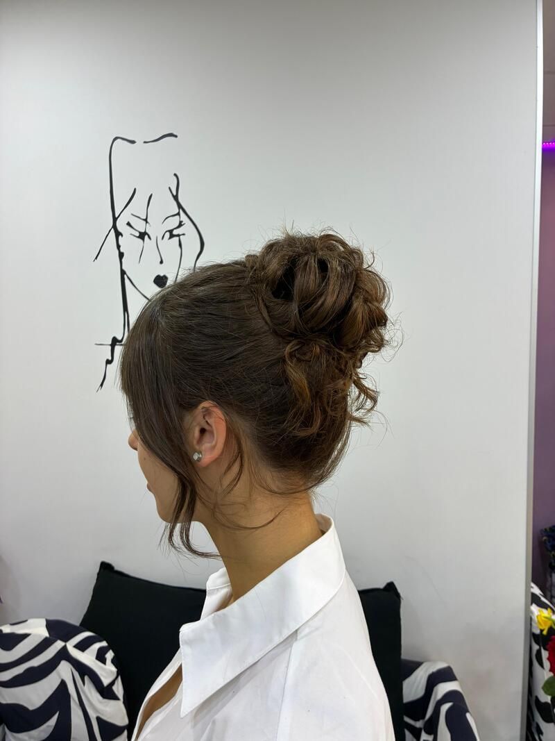 Perfil de una mujer con un moño rizado y una camisa blanca, de pie en el interior, cerca de un boceto en la pared.