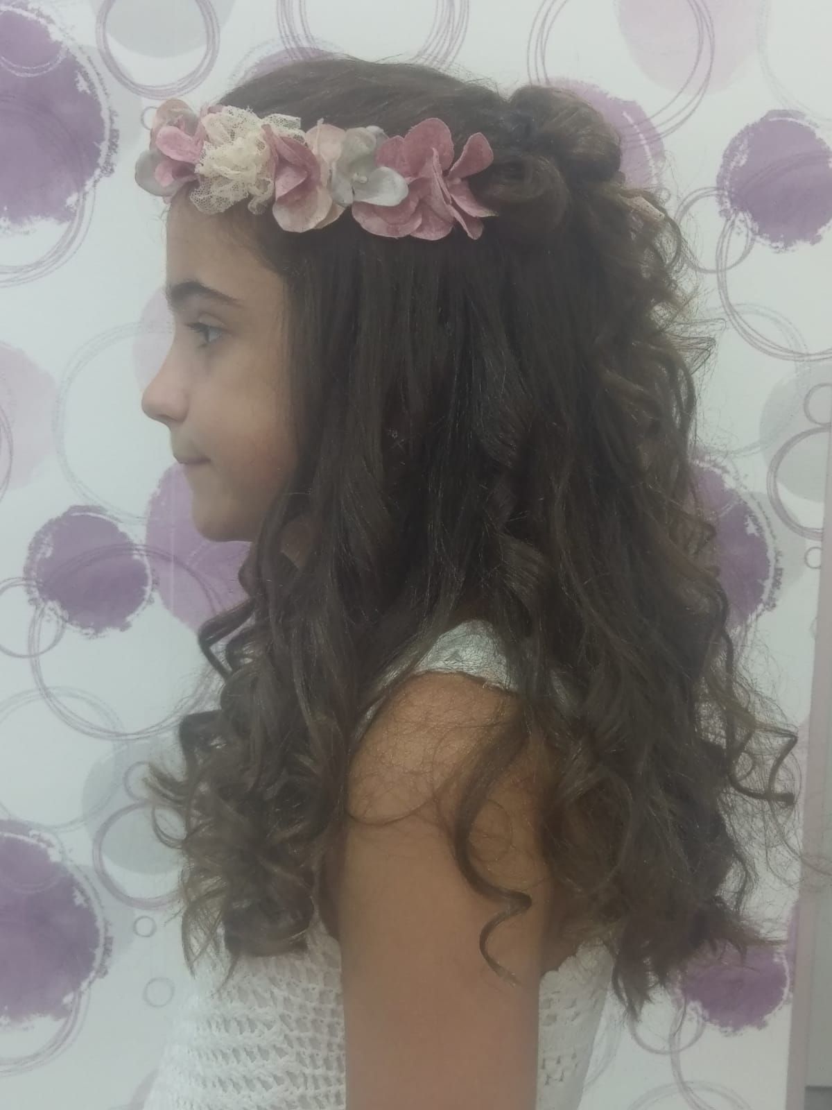 Vista de perfil de una niña con el pelo rizado y una corona de flores rosas, frente a una pared floral morada.