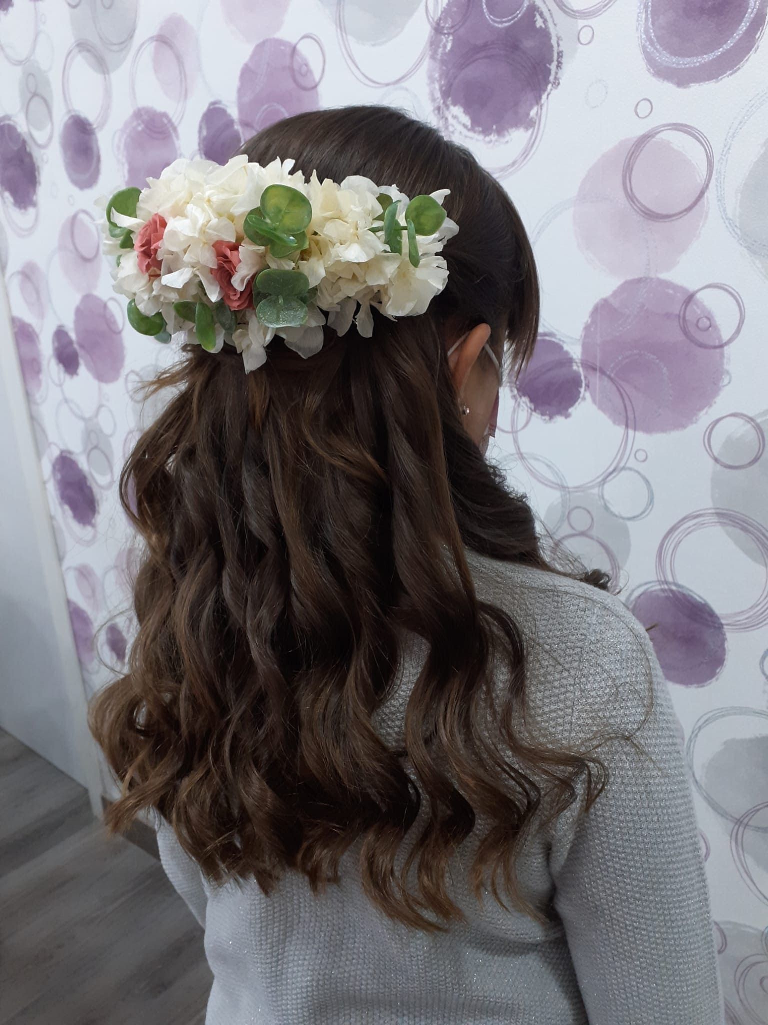 Vista de espaldas de una persona con el pelo largo y ondulado y un tocado floral blanco, frente a una pared morada estampada