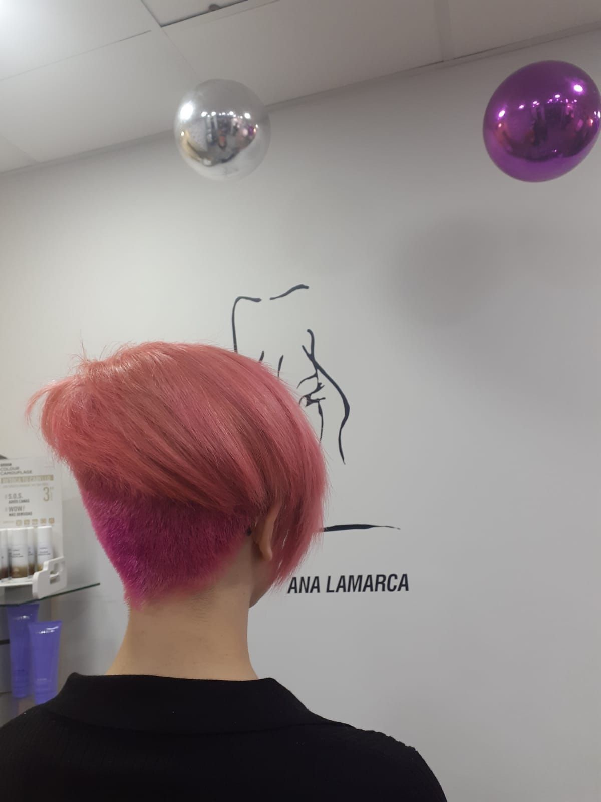 Persona de espaldas en un salón, con el pelo rosa corto y bajo globos plateados y morados.