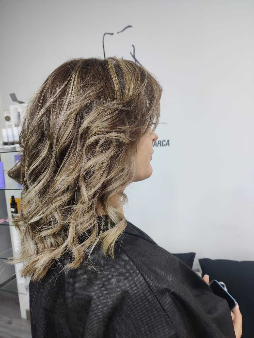 Mechas rubias en capas sobre un cabello ondulado hasta los hombros, vista lateral en un salón de peluquería