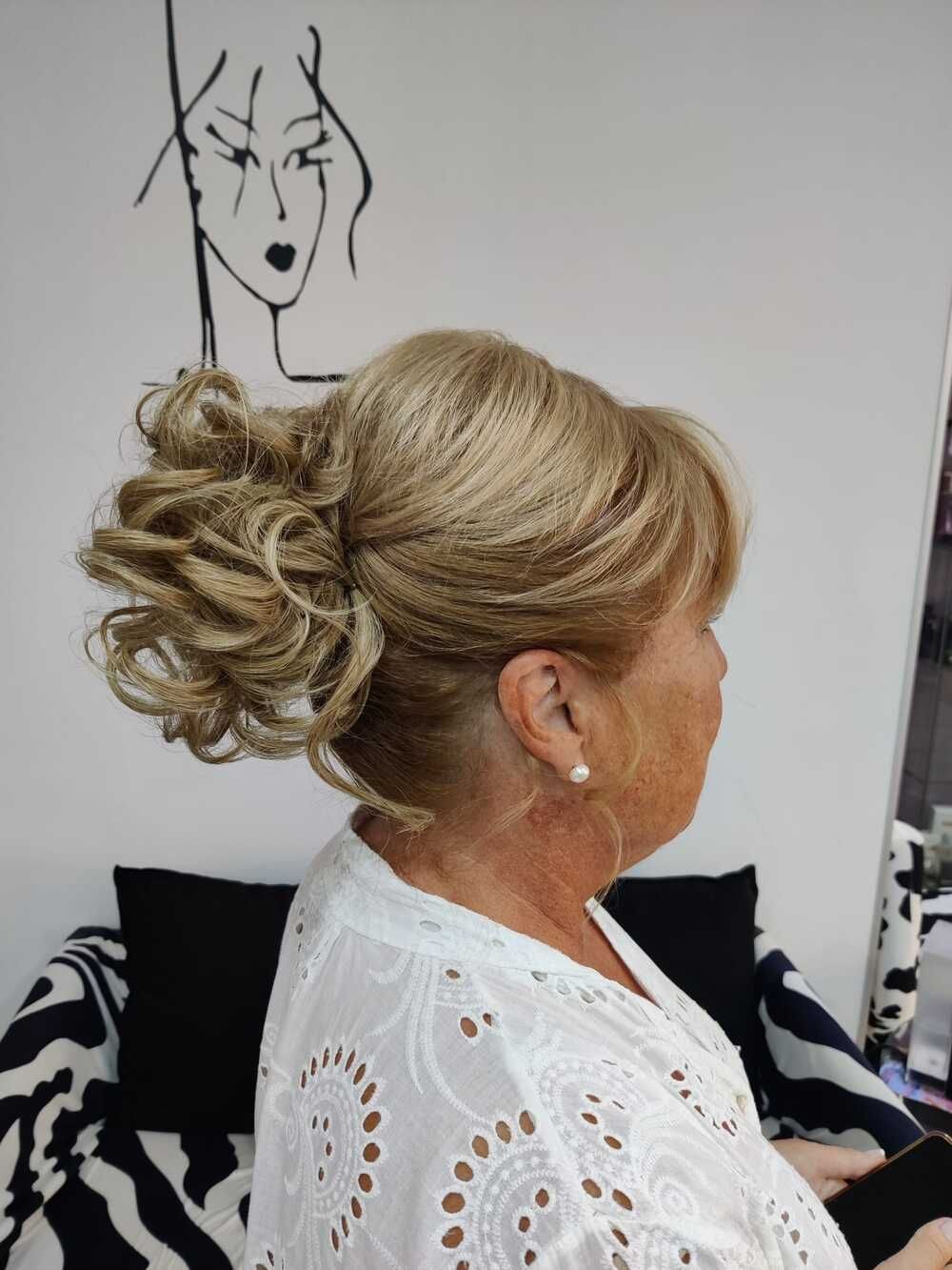 Perfil de una mujer con un elegante recogido rubio, sentada en una silla de salón y vestida con un top blanco de encaje.