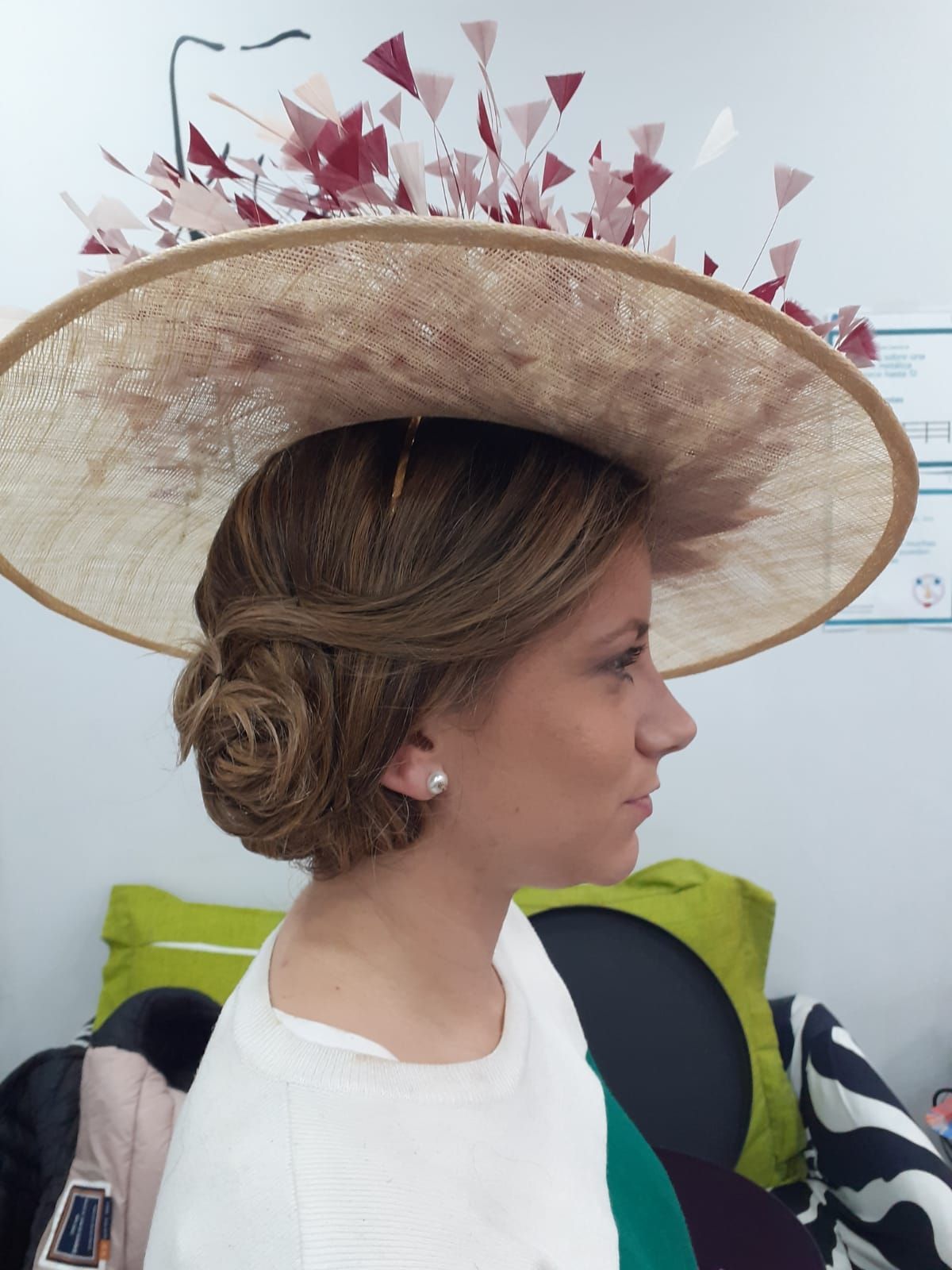 Mujer de perfil con un sombrero de paja de ala ancha y plumas rosas en un espacio interior.