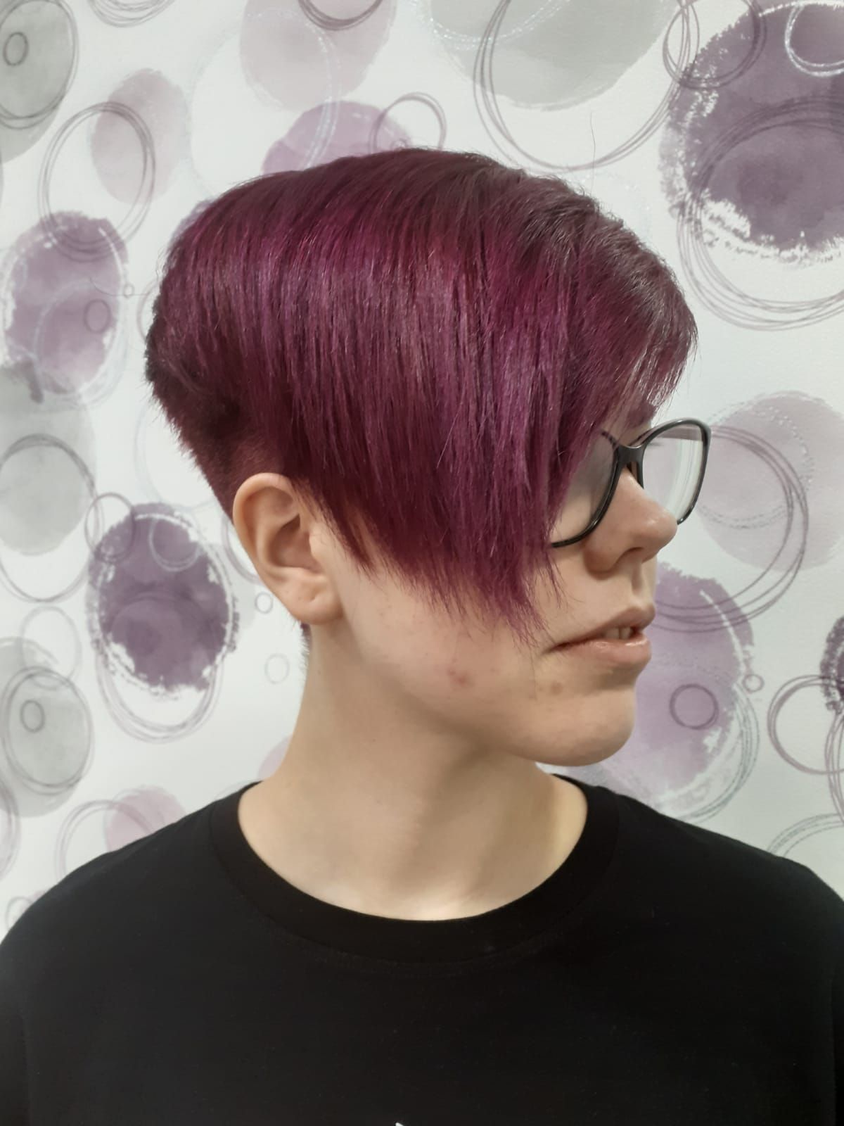 Vista de perfil de una persona con el pelo corto de color magenta y gafas, frente a una pared estampada.