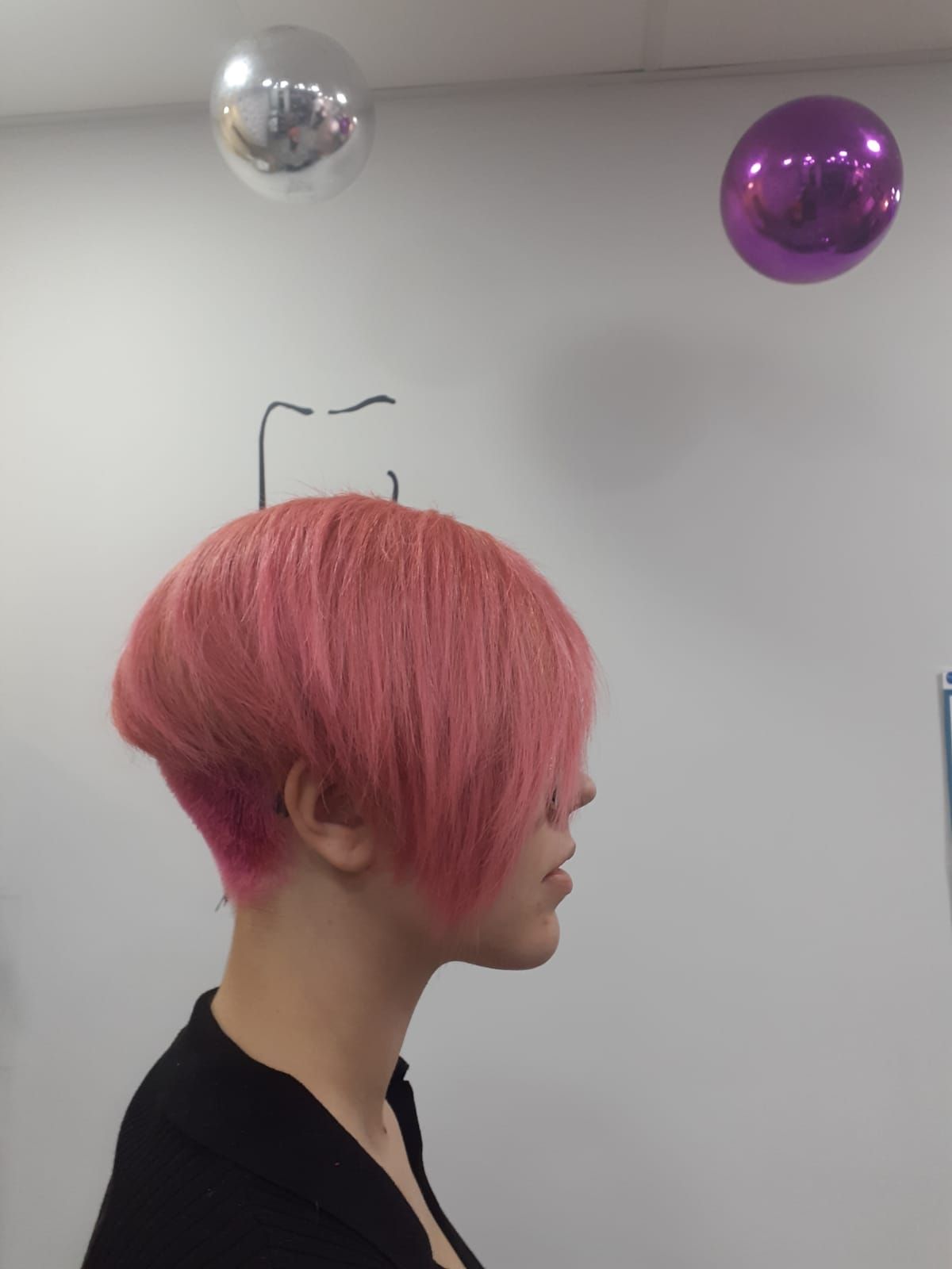 Vista de perfil de una persona con el pelo corto y rosa y una camiseta negra, en interiores, bajo unas esferas metálicas colgantes
