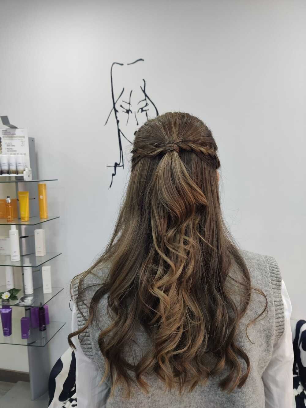 Mujer vista de espaldas, con el pelo largo, ondulado y castaño, de pie en un salón frente a un espejo de pared.