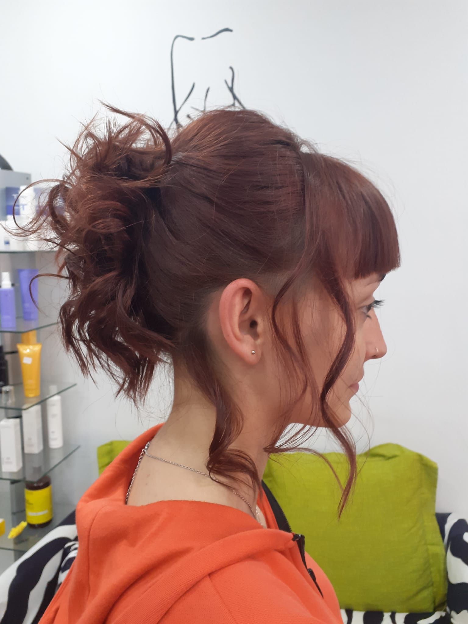 Mujer pelirroja oscura con el cabello recogido en un moño despeinado, de perfil y con una sudadera naranja con capucha en un espacio interior.